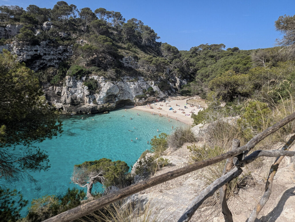10 Trendy Beaches in Europe for 2026 Pine-covered cliffs surrounding the turquoise cove of Cala Macarella, Menorca