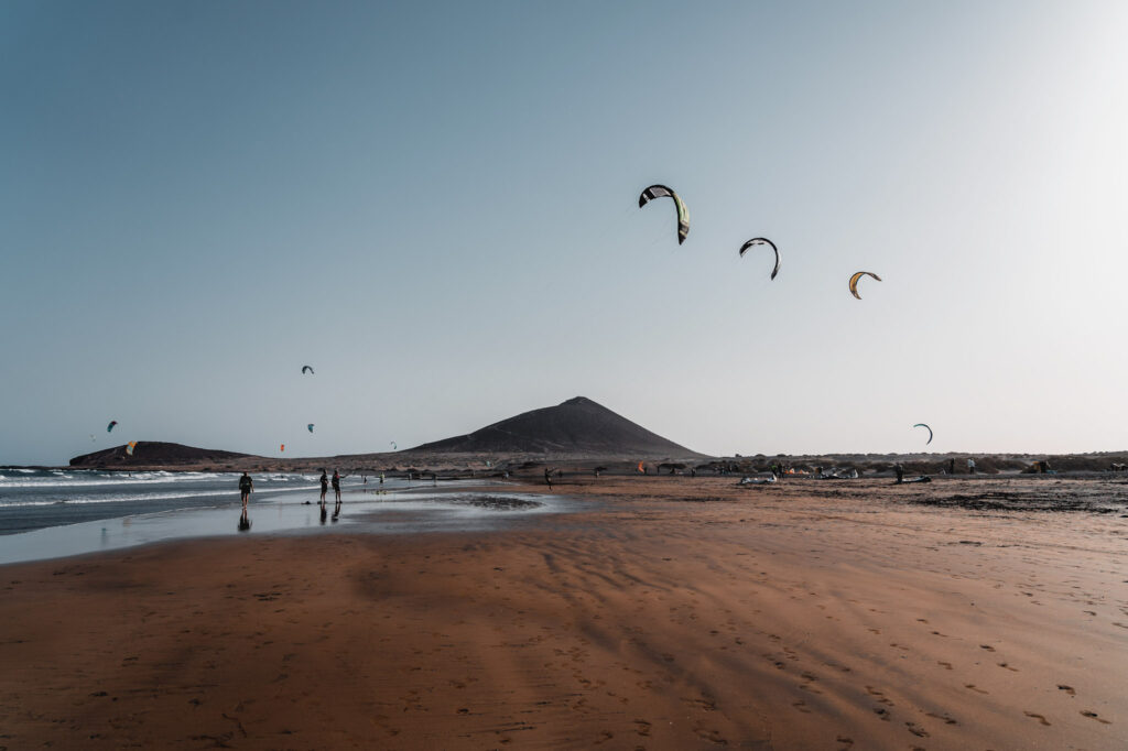 Top 10 Beaches in Tenerife Kites flying high above the wet, reddish-brown sand of Playa de El Médano beach, a famous water sports destination in Tenerife