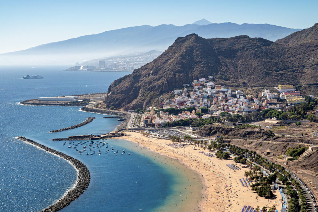 Top 10 Beaches in Tenerife Scenic overview of Playa de Las Teresitas beach in Tenerife, showing the golden shoreline, calm blue water, coastal cliffs, and a distant mountain peak