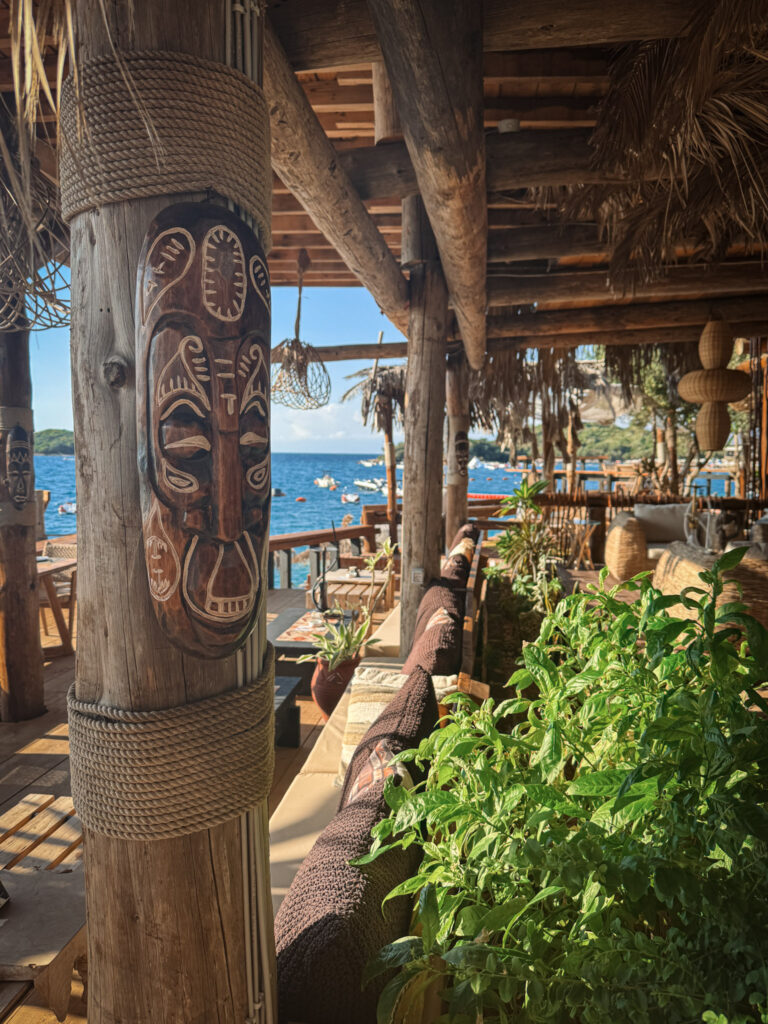 Top 10 Beaches in Ksamil, Albania An exotic tiki bar terrace at Africana Beach Club in Ksamil, Albania, featuring a carved wooden tribal mask mounted on a rope-wrapped pillar overlooking the sea.