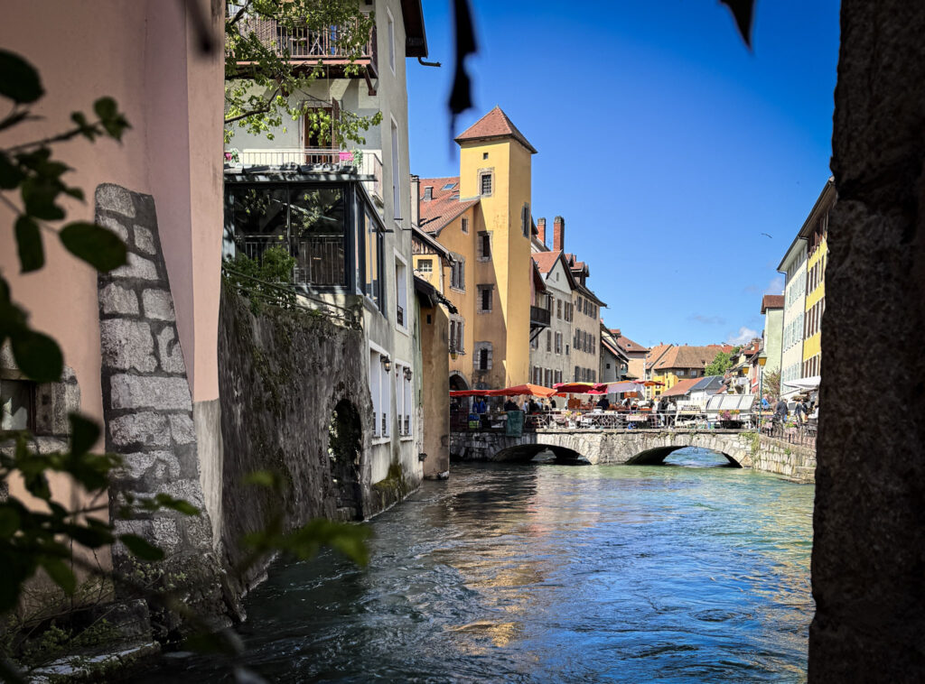 Top 10 Places in Southeastern France A scenic view of the Thiou River canal in the old town of Annecy, France, featuring historic colorful buildings, a stone bridge, and an outdoor market under a clear blue sky