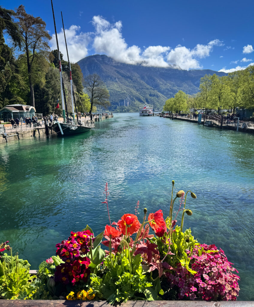 Top 10 Places in Southeastern France Vibrant red and pink flowers in a window box overlook the crystal-clear turquoise waters of the Vasse Canal leading into Lake Annecy, France, with classic boats moored along the banks and mountains under a blue sky in the background