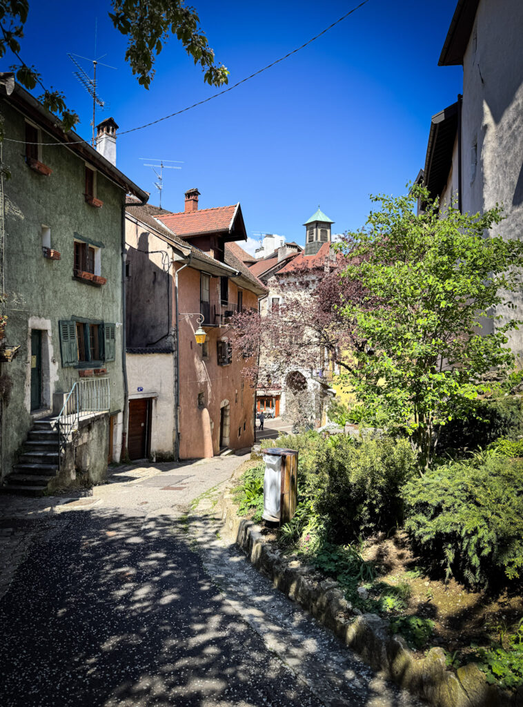 Top 10 Places in Southeastern France A cobblestone pedestrian alley in the old town of Annecy, France, lined with historic green and peach-colored stone houses, lush green gardens, and a church tower visible in the background under a blue sky