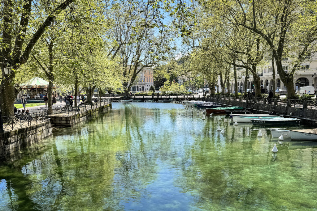 Top 10 Places in Southeastern France Small wooden rowboats are moored along the clear green waters of the Canal du Vassé, shaded by large green plane trees with a classic carousel visible in the park in Annecy, France