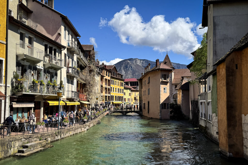 Top 10 Places in Southeastern France Outdoor restaurant patios line the clear green waters of the Thiou River canal, with pastel-colored historic buildings, a pedestrian bridge, and a distant mountain under a blue sky in Annecy, France