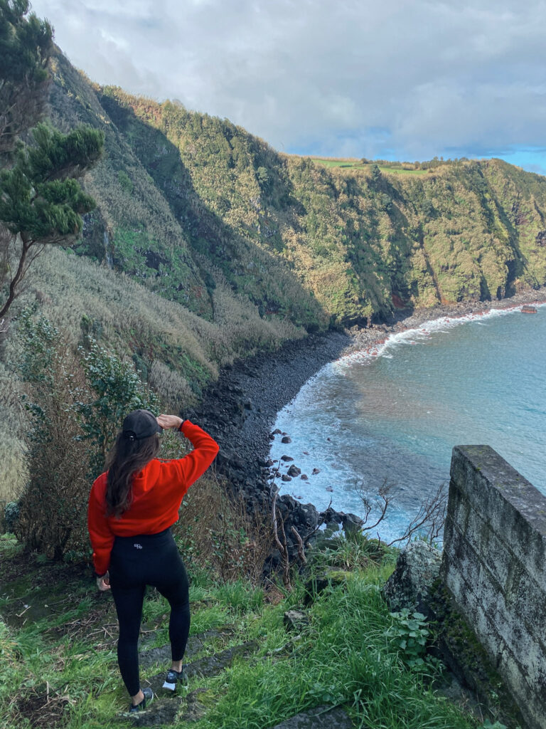 Top Things to Do in São Miguel, Azores Hiker standing on a cliffside trail overlooking a volcanic stone bay and bright blue ocean water in the Azores.