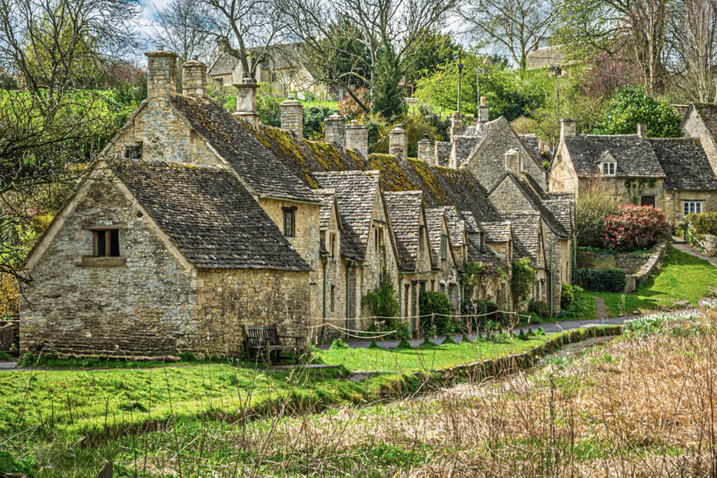Top 5 Villages in the Cotswolds A scenic view of the historic Arlington Row weavers' cottages in the village of Bibury, England. The 17th-century honey-colored stone cottages feature steeply pitched, moss-covered gabled roofs and stone chimneys, lining a narrow path next to a lush green lawn and sloping hillside under a soft, overcast sky.