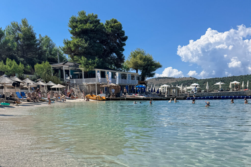 Top 10 Beaches in Ksamil, Albania The clear turquoise bay of Bora Bora Beach in Ksamil, Albania, showing swimming tourists, sunbeds, and a beachfront restaurant under a bright blue sky.