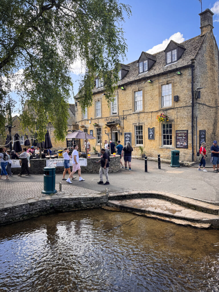 Top 5 Villages in the Cotswolds A scenic view of the historic Old Manse Hotel in Bourton-on-the-Water, England. The honey-colored stone building stands beside the shallow, flowing River Windrush, with travelers walking along the stone pathways and dining under patio umbrellas on a bright, sunny day.