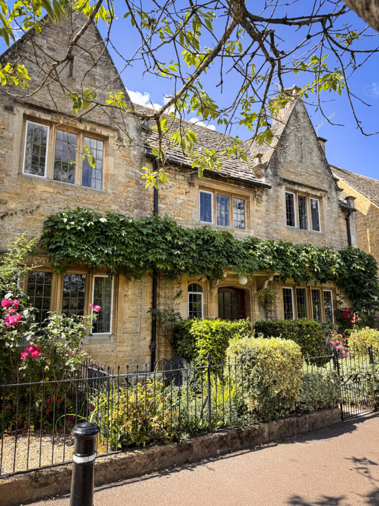 Top 5 Villages in the Cotswolds A picturesque, traditional house made of honey-colored Cotswold stone in Bourton-on-the-Water, England. The historic building features gabled roofs, stone mullioned windows, a thick layer of green ivy growing across the front facade, and a small front garden enclosed by a black wrought-iron fence filled with bright pink roses and manicured green hedges under a clear blue sky.