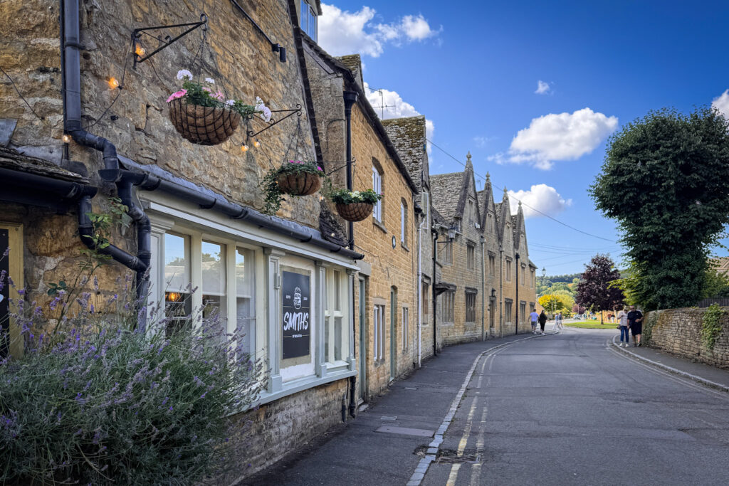 Top 5 Villages in the Cotswolds A scenic street view looking down a quiet road lined with historic honey-colored Cotswold stone buildings in Bourton-on-the-Water, England. In the foreground, the exterior of Smiths of Bourton burger restaurant features hanging flower baskets, string lights, and purple lavender bushes, with a few travelers strolling along the curving pavement under a bright blue sky with scattered clouds.