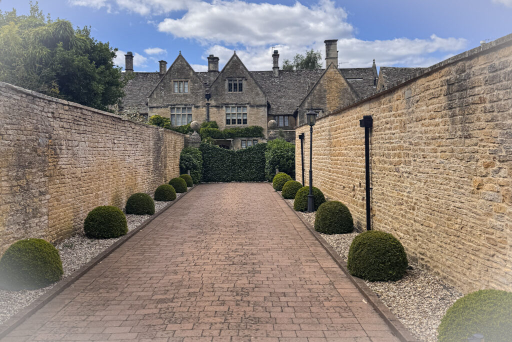 Top 5 Villages in the Cotswolds A long, brick-paved driveway flanked by tall honey-colored stone walls leads toward the historic Manor House in Bourton-on-the-Water, England. The driveway is lined with manicured, round green boxwood shrubs, a classic lamppost, and a dense green hedge gate that partially blocks the view of the grand stone mansion under a partly cloudy blue sky.