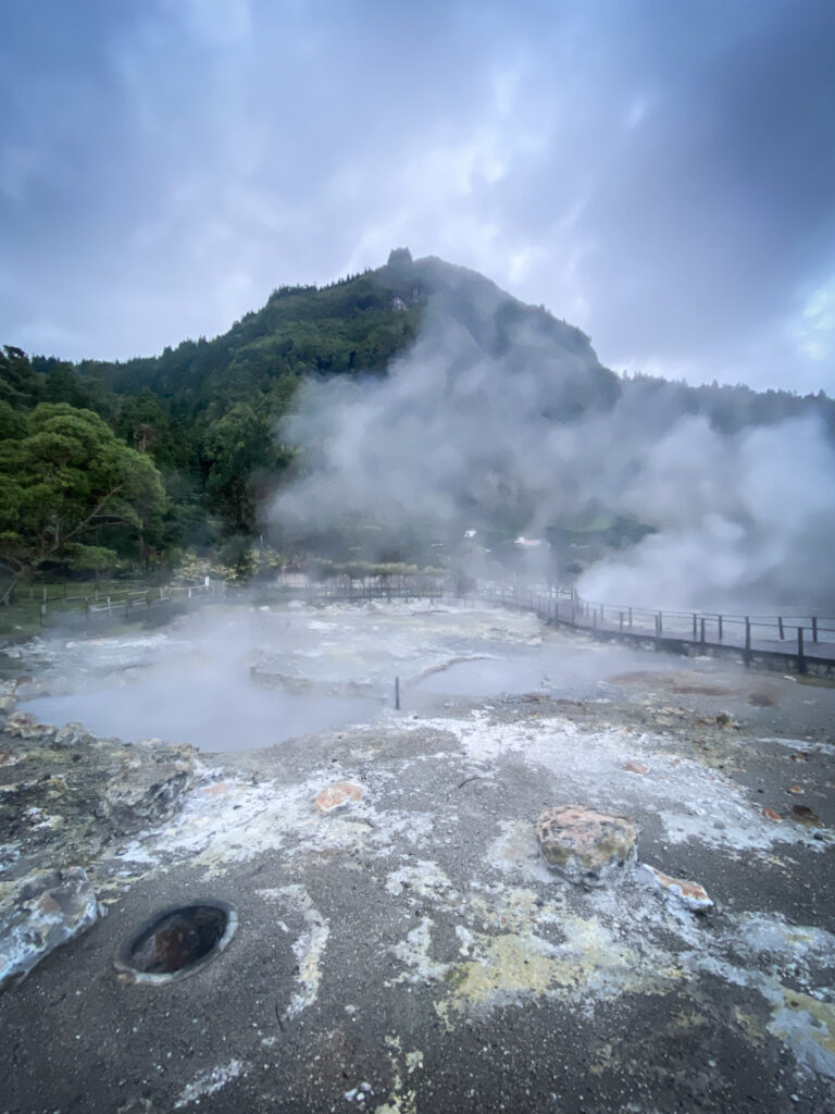 Top Things to Do in São Miguel, Azores Large clouds of sulfur steam rising from volcanic hot springs and mineral-stained ground below a prominent, lush green mountain peak under cloudy skies in Furnas, São Miguel, Azores.