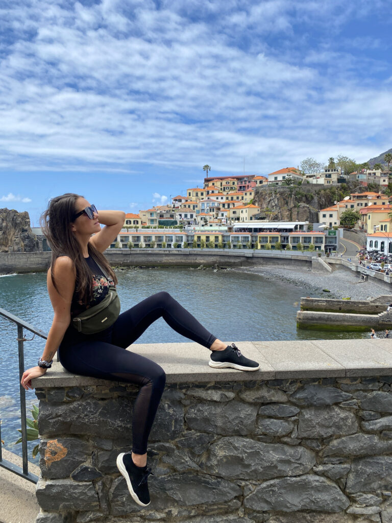 Madeira Travel Guide A traveler posing on a stone wall above a quiet ocean bay, with the terraced houses and orange roofs of Câmara de Lobos rising up the hill behind her.