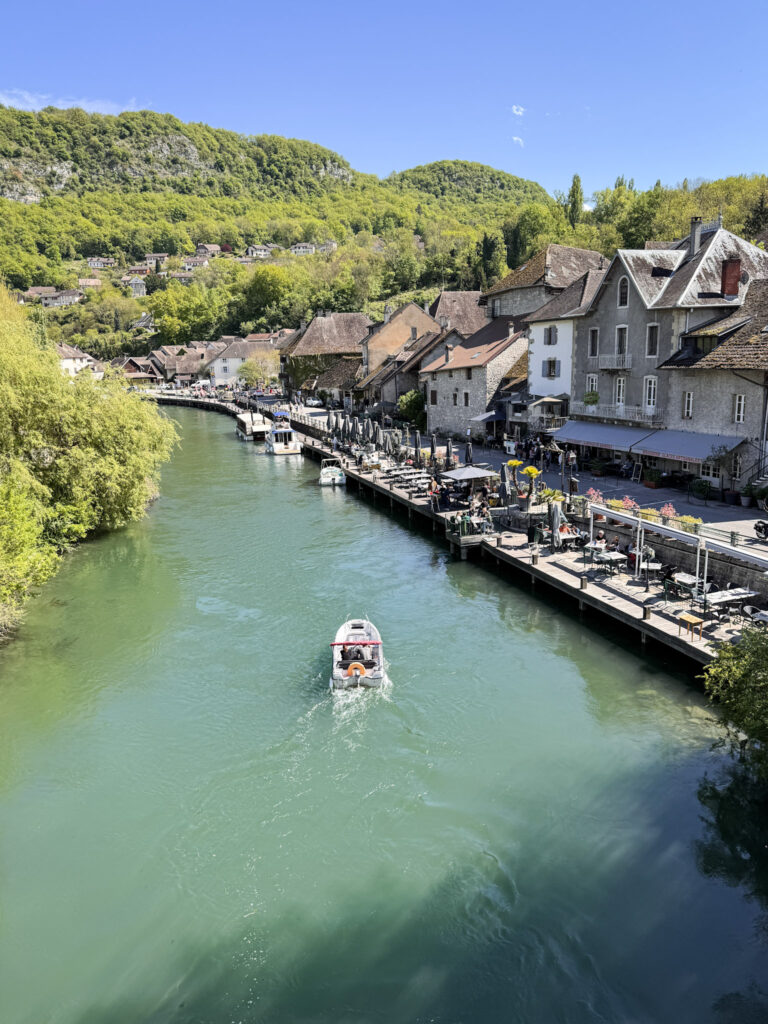 Top 10 Places in Southeastern France A small motorboat cruises down the green Canal de Savières, lined with historic stone buildings, outdoor restaurant terraces, and lush green hillsides in the village of Chanaz near Annecy, France