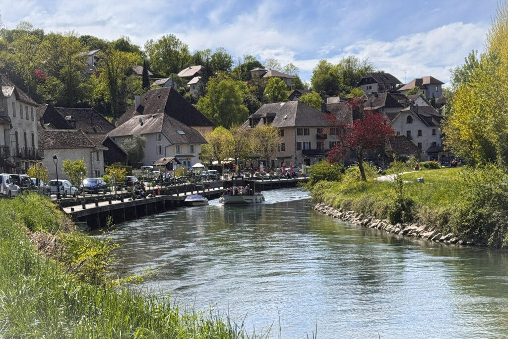 Top 10 Places in Southeastern France A small tour boat navigates a bend in the calm green Canal de Savières, with the historic stone houses of Chanaz village rising up the lush green hillside under a partly cloudy sky in France