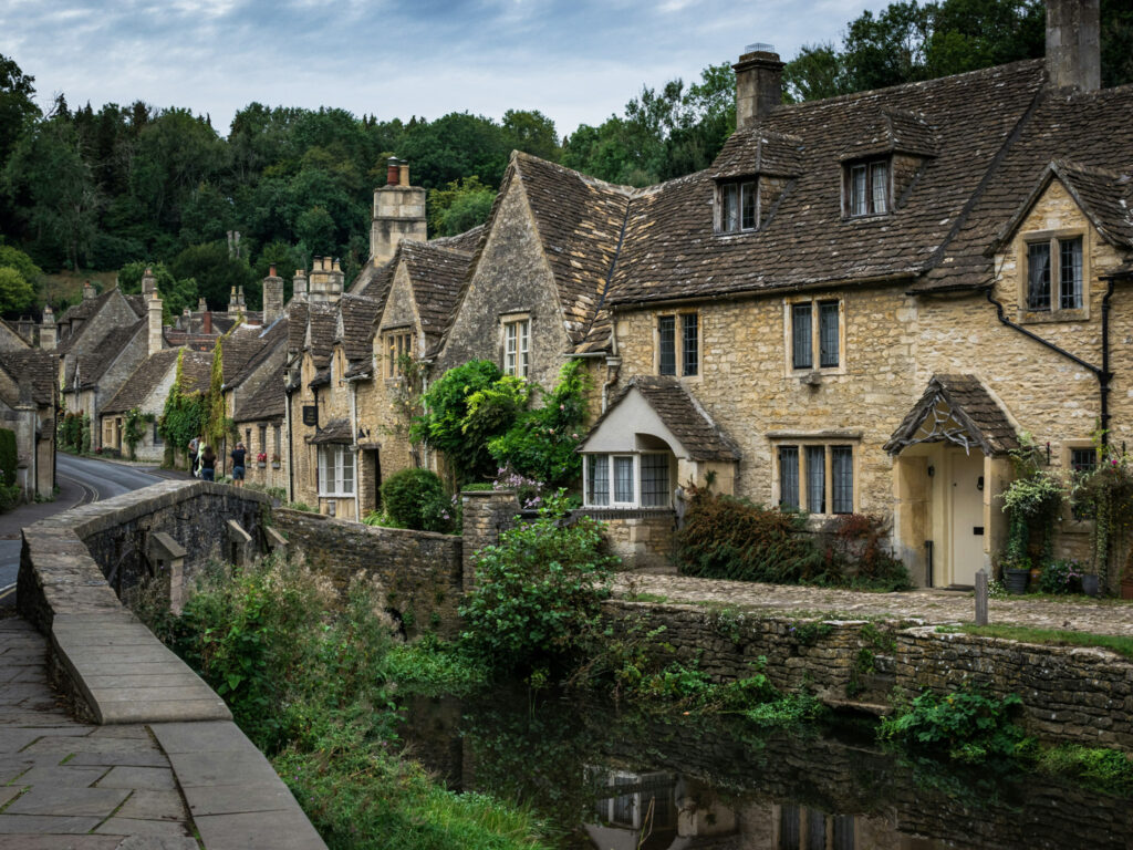 Top 5 Villages in the Cotswolds The iconic street view of Castle Combe, often called the prettiest village in England. A row of historic, honey-colored Cotswold stone cottages with steep, weathered tile roofs and dormer windows curves gently down a quiet lane. The historic limestone buildings are framed by thick green forests in the background and a low stone bridge wall in the foreground under an overcast sky.