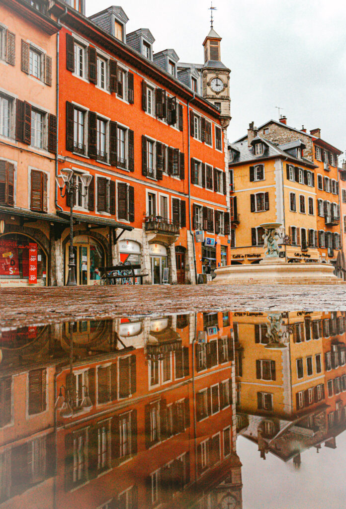 Top 10 Places in Southeastern France A clear puddle reflection on a wet cobblestone street shows bright orange and yellow historic buildings, an old clock tower, and a stone fountain in Place Notre-Dame under an overcast sky in Chambéry, France