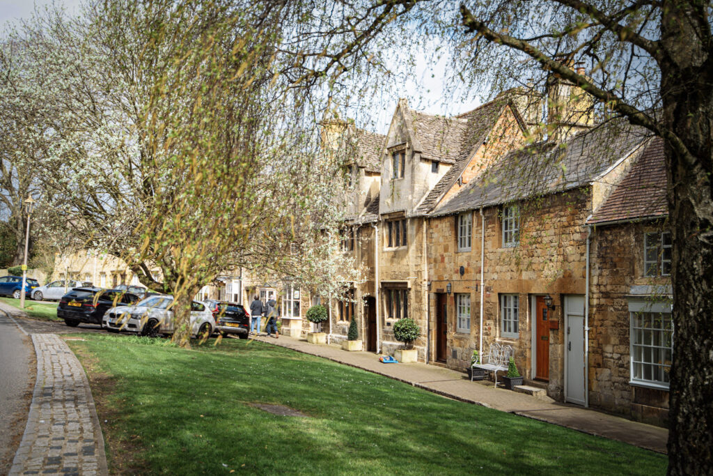 Top 5 Villages in the Cotswolds A scenic street view of historic, honey-colored Cotswold stone buildings in the market town of Chipping Campden, England. A row of multi-story limestone cottages with gabled roofs sits along a paved sidewalk, separated from a cobblestone lane by a wide, sloping green lawn. Mature trees with spring blossoms and green leaves frame the scene, with a few parked cars visible in the background under a bright sky.