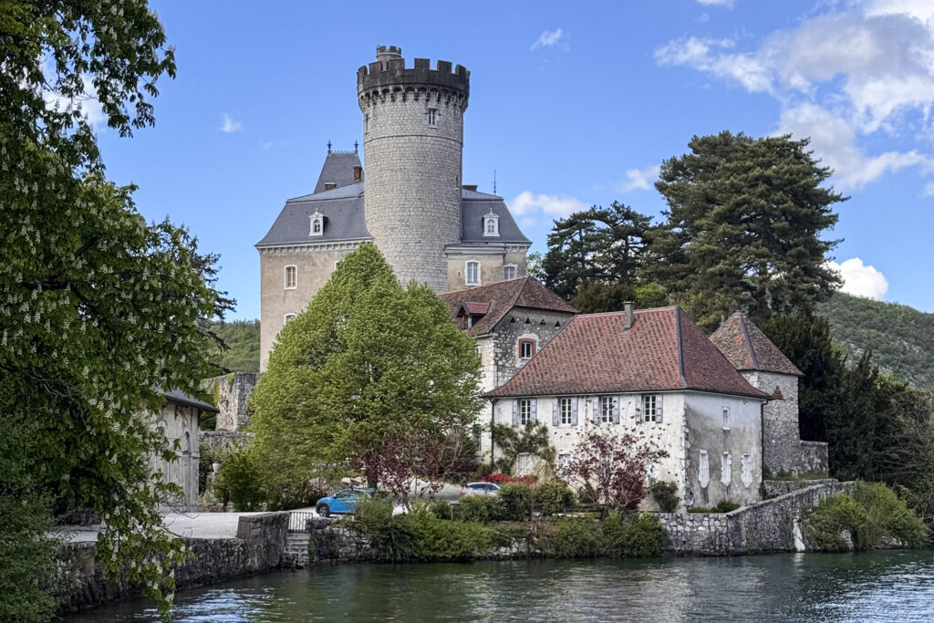 Top 10 Places in Southeastern France The medieval stone Château de Duingt with a prominent round tower sits on the edge of Lake Annecy, surrounded by green trees and old stone buildings under a blue sky in France