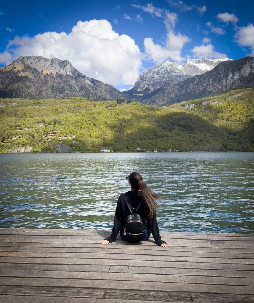 Top 10 Places in Southeastern France A traveler with a black backpack sits on a wooden dock, looking out at the calm blue water of Lake Annecy toward lush green hills and towering, snow-capped mountains under a bright blue sky in France