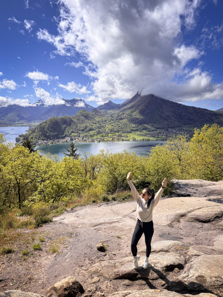 Top 10 Places in Southeastern France A female hiker stands on a rocky summit with arms raised in celebration, overlooking the panoramic blue waters of Lake Annecy, the village of Talloires, and towering mountains under a blue sky at Roc de Chère, France