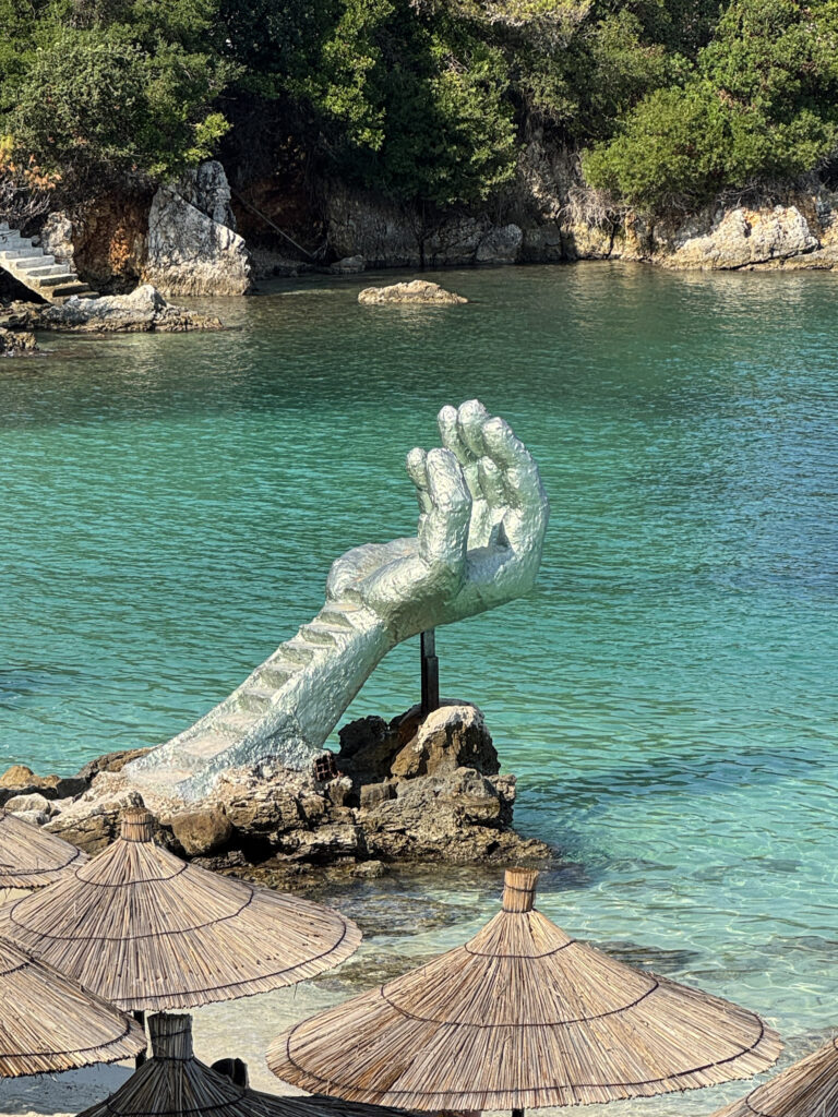 Top 10 Beaches in Ksamil, Albania The giant silver Hand of Ksamil sculpture rising from the turquoise sea past thatched straw umbrellas at Kamay Beach in Ksamil, Albania.