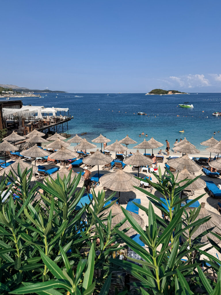 Top 10 Beaches in Ksamil, Albania Elevated view of Castle Beach next to the Hand of Ksamil in Albania, featuring thatched straw umbrellas and blue sunbeds overlooking the turquoise sea.