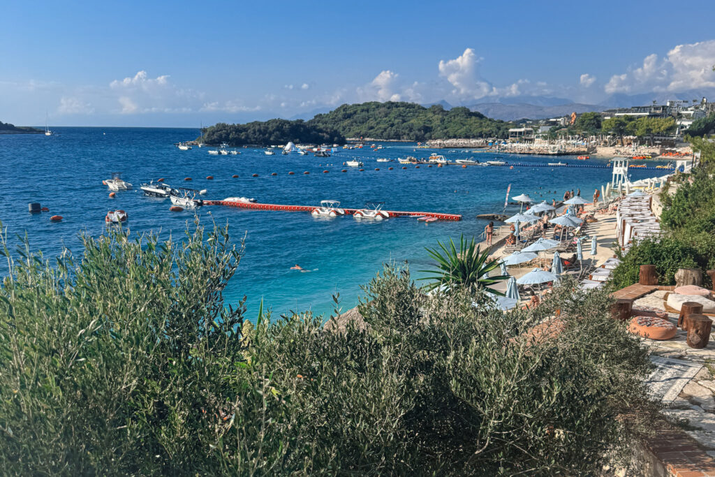 Top 10 Beaches in Ksamil, Albania Elevated panoramic view of Horizont Beach in Ksamil, Albania, showing a turquoise bay filled with boats and light blue beach umbrellas nestled along the coastline.