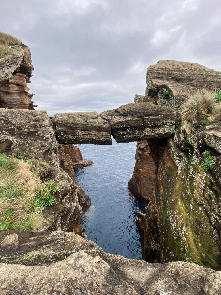 Top Things to Do in São Miguel, Azores A natural stone slab bridge connecting rugged, moss-covered volcanic sea cliffs above the deep blue ocean at the Vila Franca do Campo Islet in São Miguel, Azores.