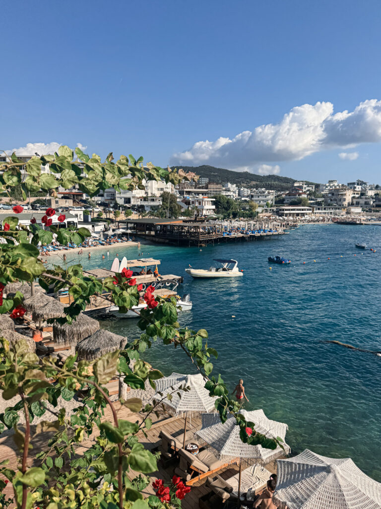 Top 10 Beaches in Ksamil, Albania Panoramic view of the main Ksamil Beach (Beach 7) in Albania, showing boats anchored in a blue ocean bay lined with hotels and red bougainvillea flowers.