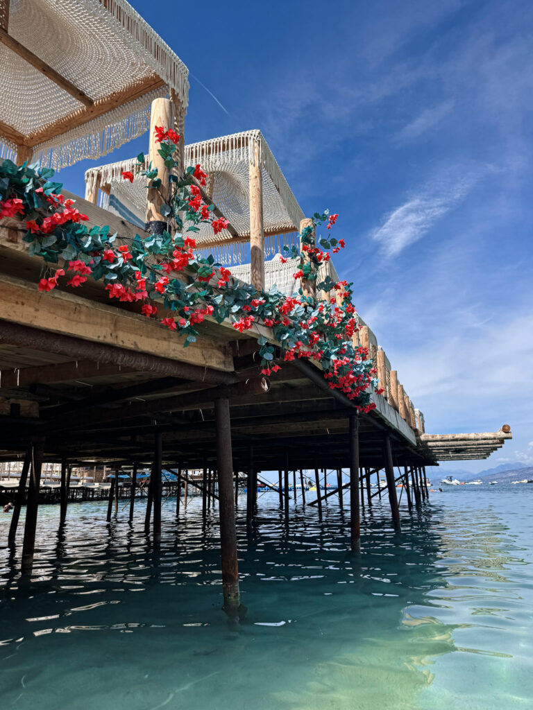Top 10 Beaches in Ksamil, Albania A wooden overwater pier decorated with vibrant red flowers and shaded cabanas overlooking the clear blue sea in Ksamil, Albania.