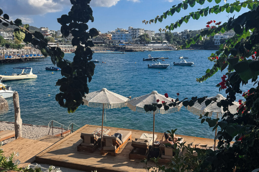 Top 10 Beaches in Ksamil, Albania Elevated view of Africana Beach Club in Ksamil, Albania, showing a wooden sun deck with luxury sunbeds and fringed umbrellas overlooking a blue bay filled with boats.