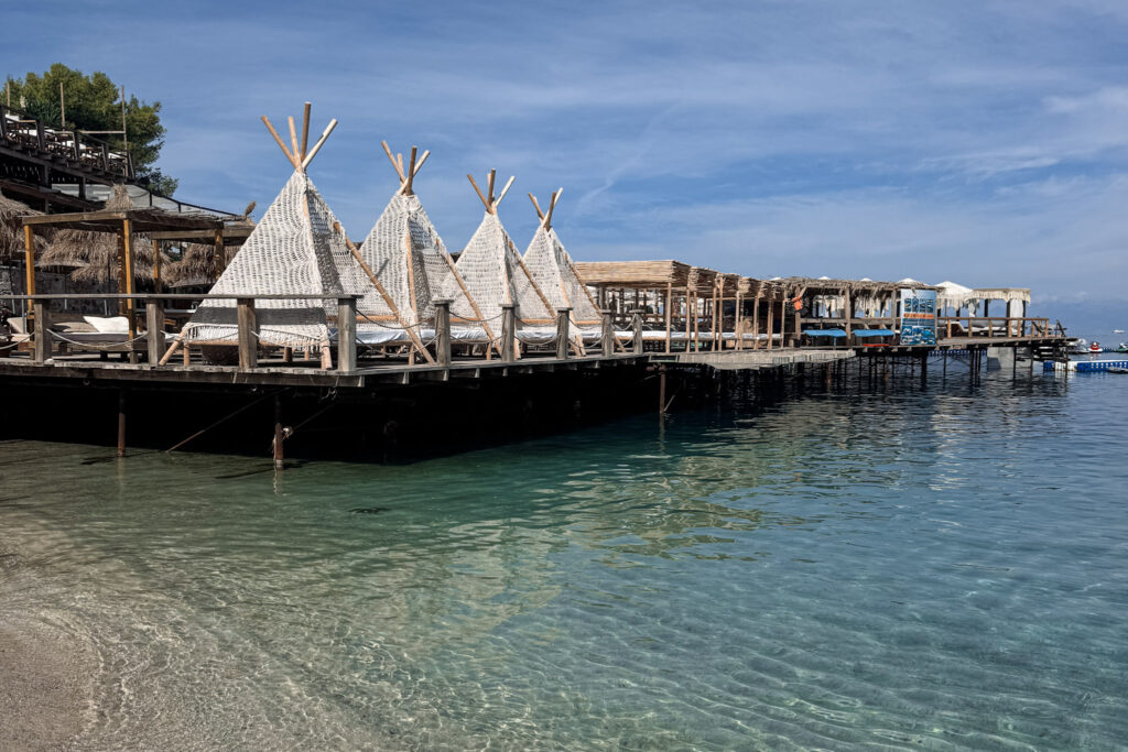 Top 10 Beaches in Ksamil, Albania Teepee-style beach cabanas on a wooden overwater deck next to crystal-clear turquoise water in Ksamil, Albania.
