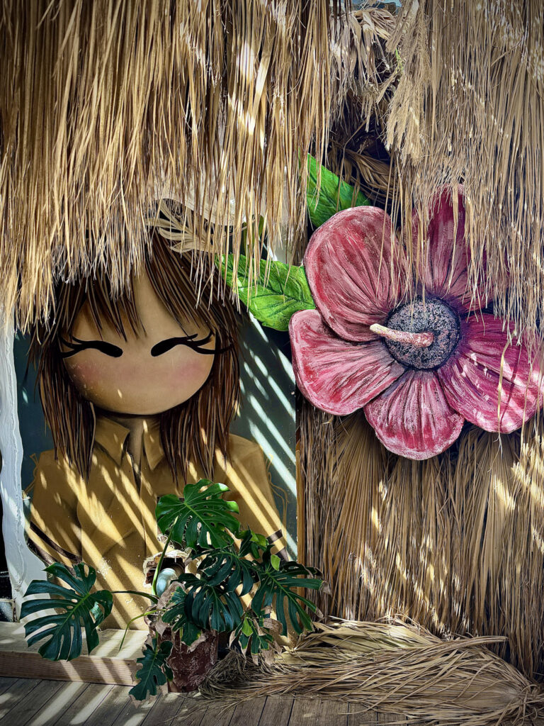 Top 10 Beaches in Ksamil, Albania Tropical tiki-style beach club decor with a large stylized doll character and a giant pink hibiscus flower under a thatched straw roof in Ksamil, Albania.