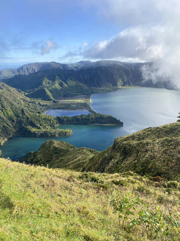 Top Things to Do in São Miguel, Azores Panoramic view of Lagoa do Fogo crater lake surrounded by steep, lush green volcanic mountains and low clouds on a sunny day in São Miguel, Azores.