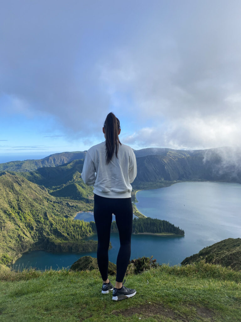 Top Things to Do in São Miguel, Azores Hiker standing on a grassy ridge overlooking a massive crater lake surrounded by green mountains in the Azores.