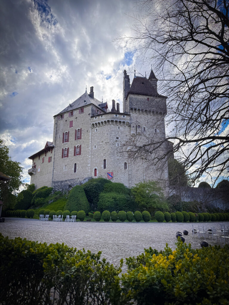 Top 10 Places in Southeastern France The imposing stone courtyard facade of the medieval Château de Menthon-Saint-Bernard featuring red-shuttered windows, rounded turrets, and manicured green hedges under a dramatic cloudy sky in France