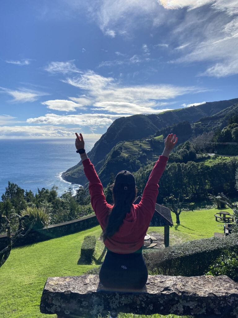 Top Things to Do in São Miguel, Azores Hiker with raised arms overlooking a coastal park and green mountains by the ocean in the Azores.