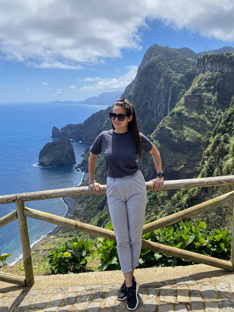 Madeira Travel Guide A traveler posing at the Miradouro da Rocha do Navio viewpoint, with towering lush green volcanic cliffs, sea stacks, and a waterfall plunging into the blue ocean in Santana, Madeira.