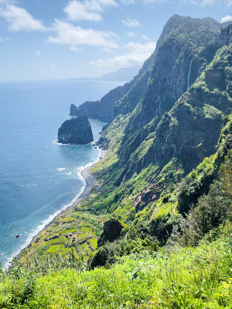 Madeira Travel Guide A sweeping clifftop view from Miradouro da Rocha do Navio, showcasing towering, lush green volcanic cliffs, a thin line of pebble beach below, and massive rocky sea stacks rising from a bright blue sea.