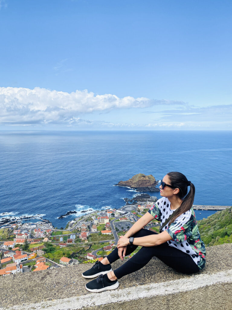 Madeira Travel Guide Traveler sitting at the Miradouro da Santinha viewpoint, looking over a coastal town and the blue ocean in Madeira.