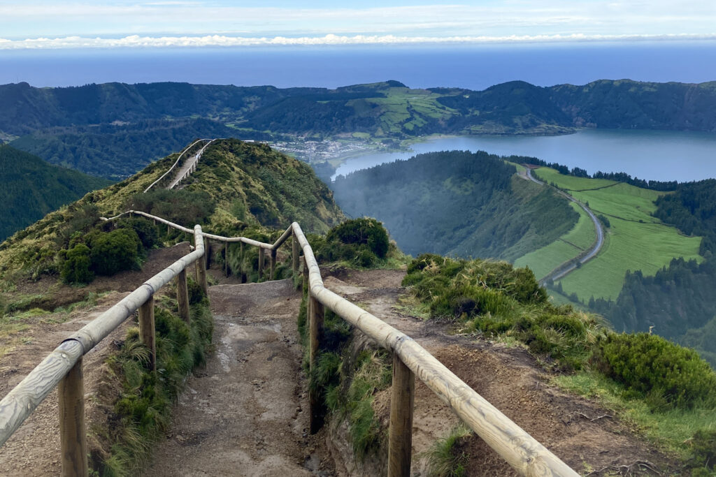 Top Things to Do in São Miguel, Azores Narrow dirt hiking path with wooden handrails leading down a steep ridge towards the volcanic lakes and green caldera of Sete Cidades, São Miguel, Azores.
