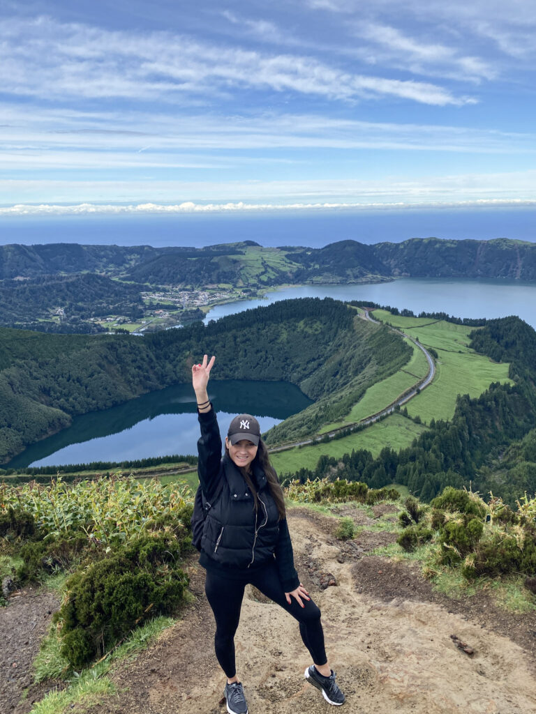 Top Things to Do in São Miguel, Azores A hiker in black athletic wear flashing a peace sign on a mountain ridge overlooking the deep blue Lagoa do Santiago and Sete Cidades lakes in São Miguel, Azores.