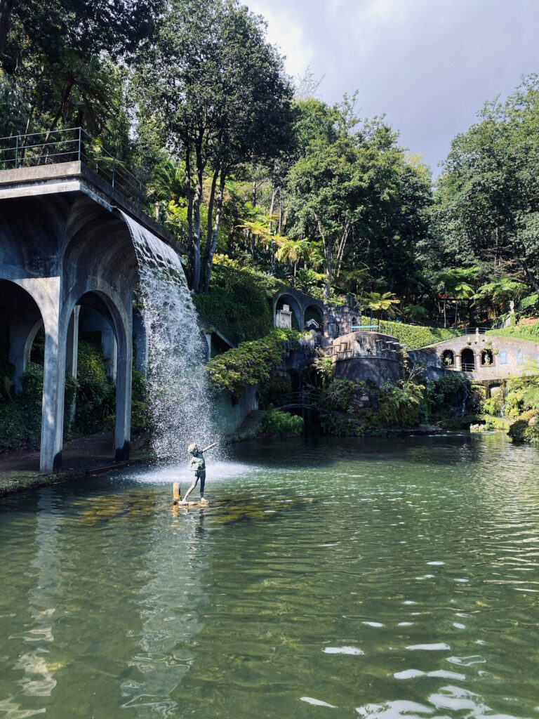 Madeira Travel Guide A massive artificial waterfall cascading from a stone arched structure into a green lake, with a statue in the center at Monte Palace Tropical Garden.