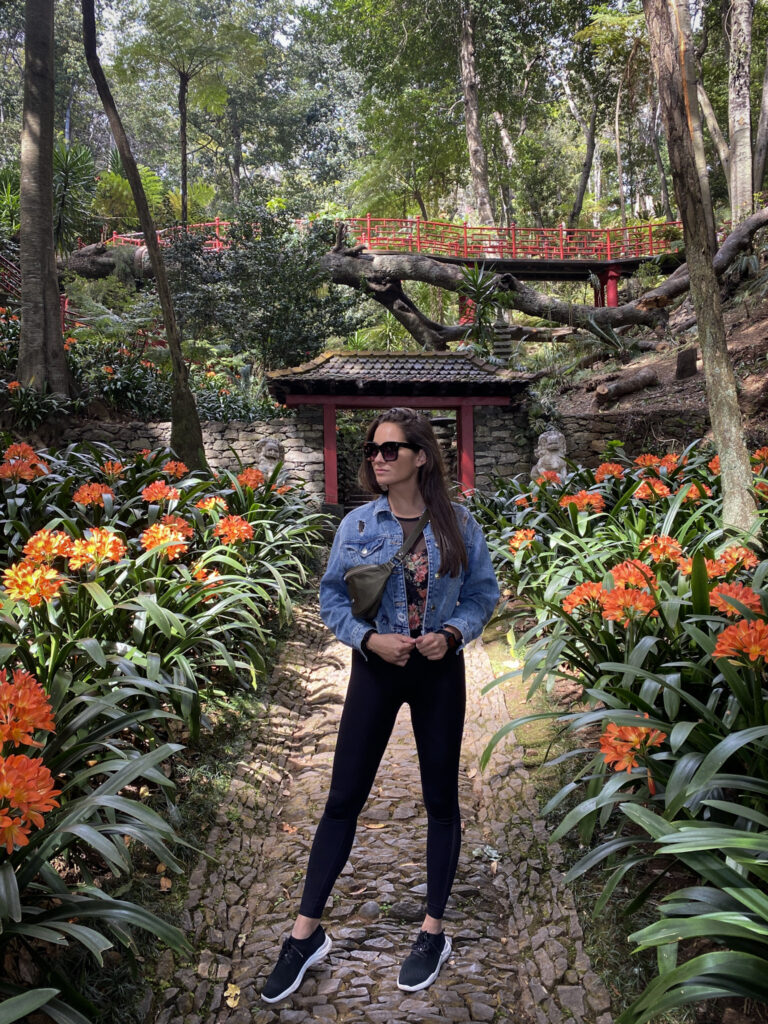Madeira Travel Guide A traveler posing on a cobblestone walkway surrounded by lush green foliage and blooming orange clivia flowers, leading toward a traditional stone gate and a red garden bridge.
