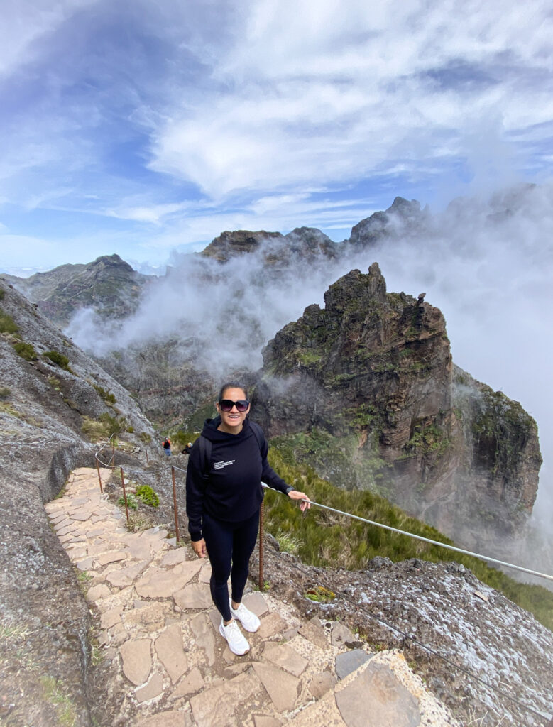 Madeira Travel Guide A traveler posing on the stone-paved hiking trail of PR1 Vereda do Arieiro in Madeira, with massive, jagged volcanic peaks and rocky ridges partially shrouded by dramatic white mountain fog and clouds.