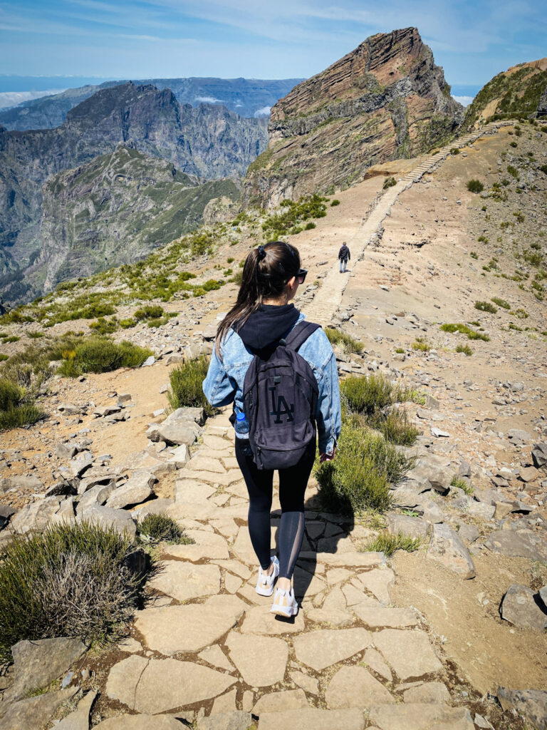 Madeira Travel Guide A traveler with a backpack seen from behind as she hikes down a paved stone ridge path along the PR1 Vereda do Arieiro trail, surrounded by the dramatic, high-altitude volcanic peaks and sweeping valley vistas of Madeira.