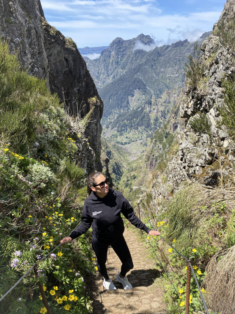 Madeira Travel Guide A traveler posing on a stone-paved section of the PR1 Vereda do Arieiro trail, framed by steep volcanic rock cliffs, yellow wildflowers, and an expansive backdrop of a lush green valley and the sharp peaks of Pico do Arieiro in Madeira.