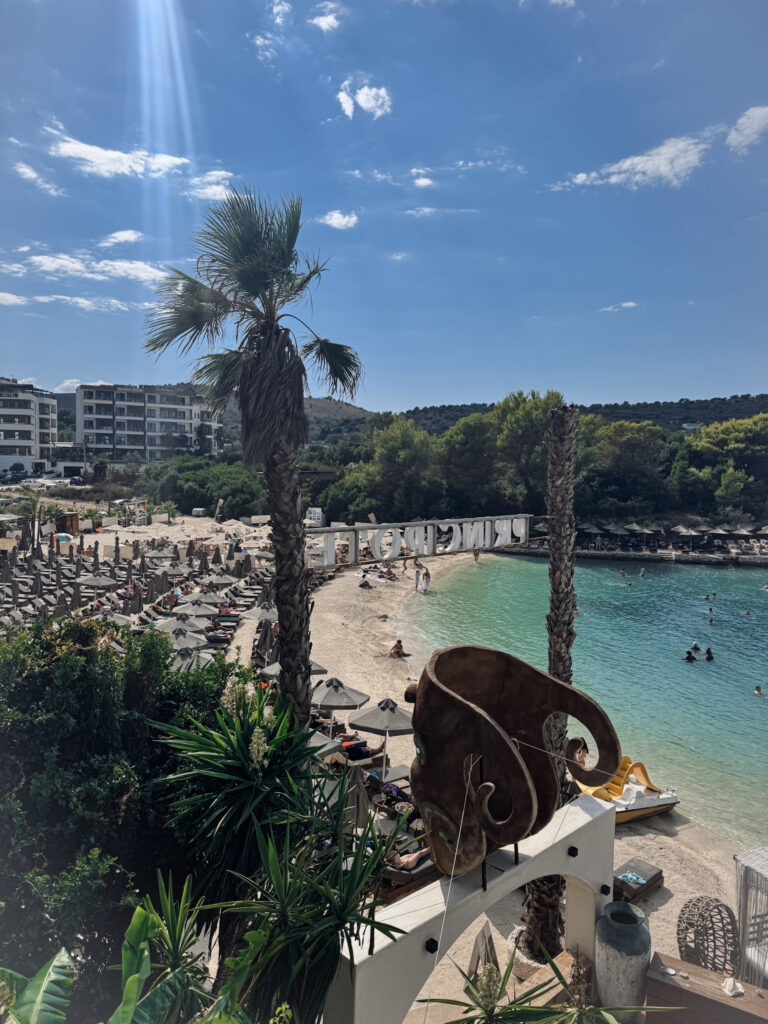 Top 10 Beaches in Ksamil, Albania Elevated panoramic view of Paradise Beach in Ksamil, Albania, showing a turquoise bay with a sandy beach, palm trees, and coastal hotels.